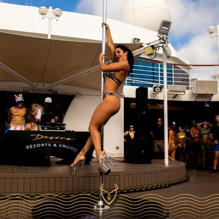 A woman in a sparkling bikini and high heels performs a pole dance on the deck of a Desire Resorts & Cruises ship. A DJ and dancers in gold outfits entertain the crowd in the background, while onlookers in swimwear gather around. The scene exudes a bold, party atmosphere with an upscale, adults-only vibe.