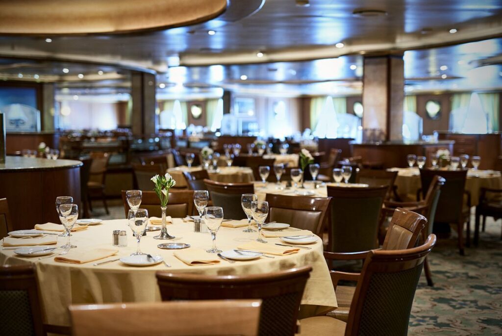 This image features the dining room aboard a Princess cruise ship, showcasing an inviting space with neatly arranged tables covered in cream-colored tablecloths. The setup includes polished glassware, cutlery, and floral centerpieces, creating an elegant atmosphere. The warm lighting and spacious layout add to the sophisticated dining experience for guests.