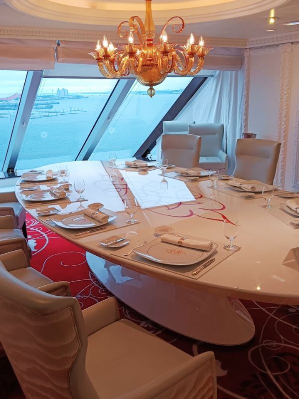 This image shows an elegant dining room on the Oceania Mariner cruise ship. A large, glossy white oval table is set with fine dining cutlery and plates, surrounded by plush high-backed chairs. The room is accentuated by a grand golden chandelier hanging from the ceiling, casting a warm glow over the table. Floor-to-ceiling windows offer stunning views of the ocean and distant harbor, creating a luxurious atmosphere for an upscale dining experience. The red carpet adds a pop of color to the otherwise neutral and sophisticated space.
