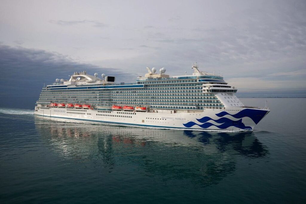 The image is of the Discovery Princess cruise ship, elegantly gliding through calm waters. The ship's sleek white and blue hull, adorned with the distinctive Princess Cruises' wave design, reflects a clear sky. The multiple decks and rows of balconies on the ship indicate a large, modern vessel, offering a variety of amenities and accommodations, ready to provide its guests with an exceptional cruising experience.