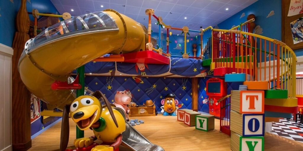 This image shows a vibrant, Toy Story-themed indoor play area featuring a large yellow spiral slide, colorful climbing structures, and oversized blocks spelling “TOY.” Prominent characters like Slinky Dog, Hamm, and Mr. Potato Head are part of the decor, making it a playful, immersive space for young children. The background is decorated with stars, rockets, and space motifs to match the movie's theme.