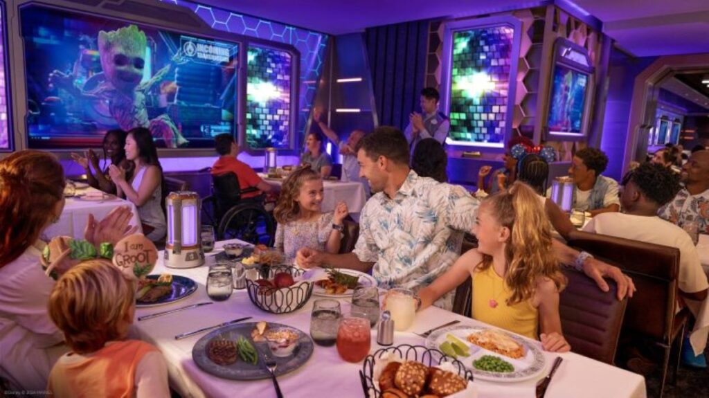 This image features a lively Marvel-themed dining experience on Disney Treasure. Families enjoy their meals at elegantly set tables while surrounded by futuristic decor, complete with glowing panels and displays featuring beloved Marvel characters like Groot. The atmosphere combines fine dining with immersive entertainment, creating a magical and engaging experience for guests of all ages.