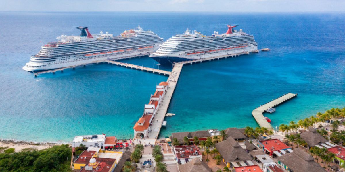Aerial view of two large cruise ships docked at a tropical pier, with striking turquoise waters surrounding them. Nearby, a cluster of colorful buildings with thatched roofs creates a picturesque scene that captures the essence of a vibrant vacation destination.