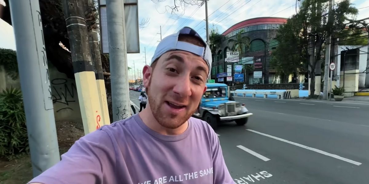 Drew Binsky stands on a sidewalk in the Philippines, smiling at the camera while wearing a lavender shirt and a backwards cap. Behind him, a traditional Filipino jeepney drives down the road near a commercial building with multiple signs and arched windows.