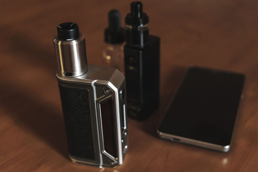 A close-up of vaping paraphernalia on a wooden surface: an e-cigarette with a dripping atomizer stands beside a bottle of vape liquid, with a smartphone lying nearby, illustrating a modern smoke-free lifestyle choice or a vaping session.