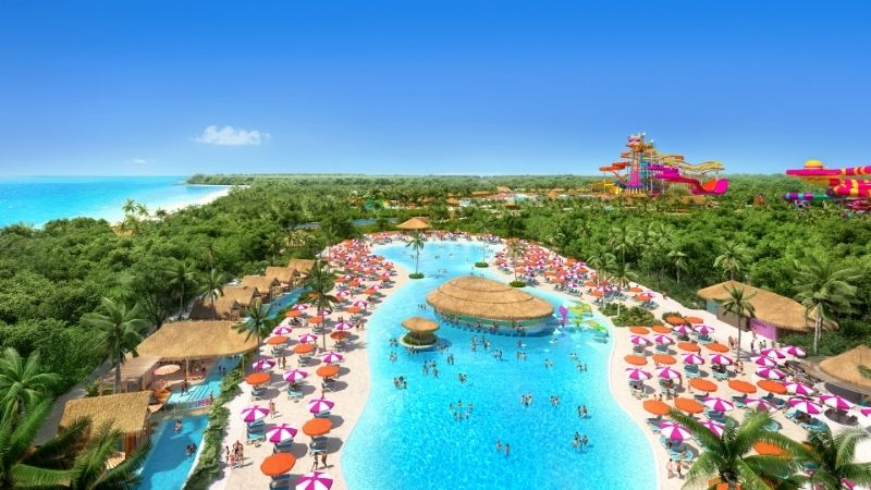 This image shows El Hideaway, the adults-only neighborhood at Royal Caribbean’s Perfect Day at Costa Maya, Mexico. The serene lagoon-style pool is lined with pink and purple umbrellas, thatched-roof cabanas, and tropical greenery, with a large swim-up bar at its center and a view of the bold water slides in the distance.