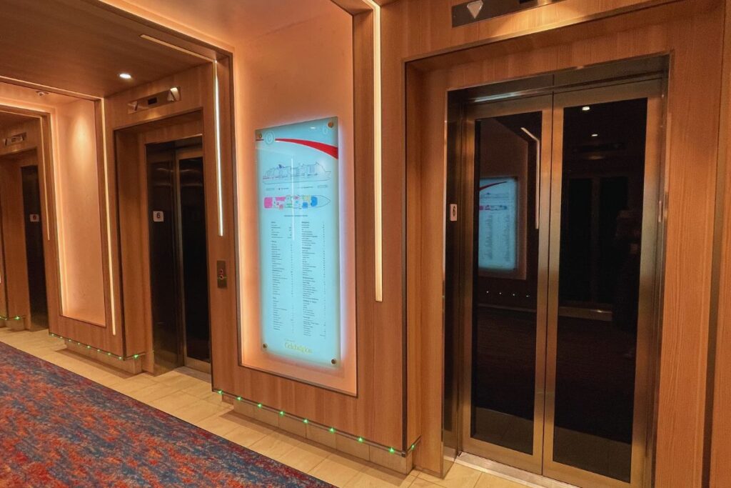 A modern elevator lobby on a Carnival cruise ship, featuring two elevator doors with wooden paneling and illuminated floor indicators. A detailed ship map is mounted between the elevators, assisting passengers in navigating the different decks. The lobby is warmly lit, with a colorful carpet enhancing the inviting atmosphere.