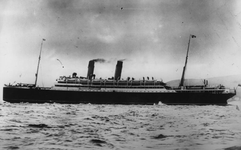 This black and white photograph shows a large early 20th-century ocean liner sailing through choppy waters, with two smoke stacks emitting dark smoke into a cloudy sky. The ship appears to be a passenger or mail steamer, featuring multiple decks and lifeboats, with flags flying at both bow and stern.