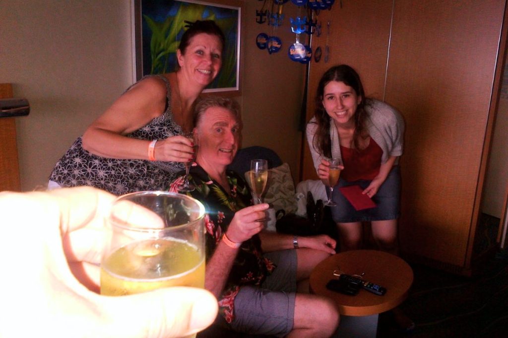 A family celebration with champagne on board Oasis of the Seas, featuring a man in a floral shirt, two smiling women, and a close-up of a hand holding a drink, set against a cabin backdrop with vibrant artwork.