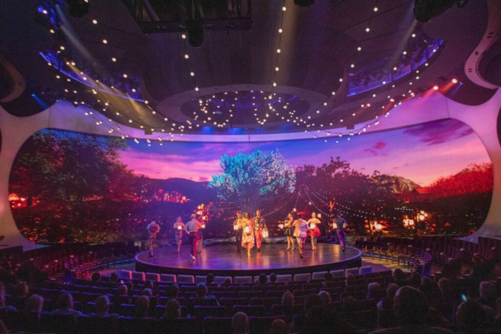 This image showcases an entertainment performance aboard the Celebrity Apex, taking place on a grand, futuristic stage. The venue features a massive curved LED screen displaying a vivid, colorful backdrop of a twilight scene, enhancing the immersive experience. Performers in vibrant costumes are in the spotlight, surrounded by a captivated audience seated in a state-of-the-art theater designed for world-class shows.