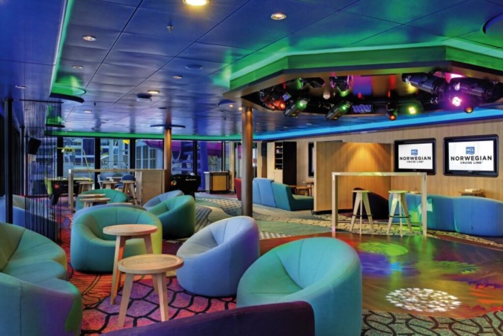 The vibrant and modern Entourage teen lounge on Norwegian Epic, featuring cozy seating, colorful lighting, and multiple screens for entertainment, designed as a fun hangout space for younger passengers