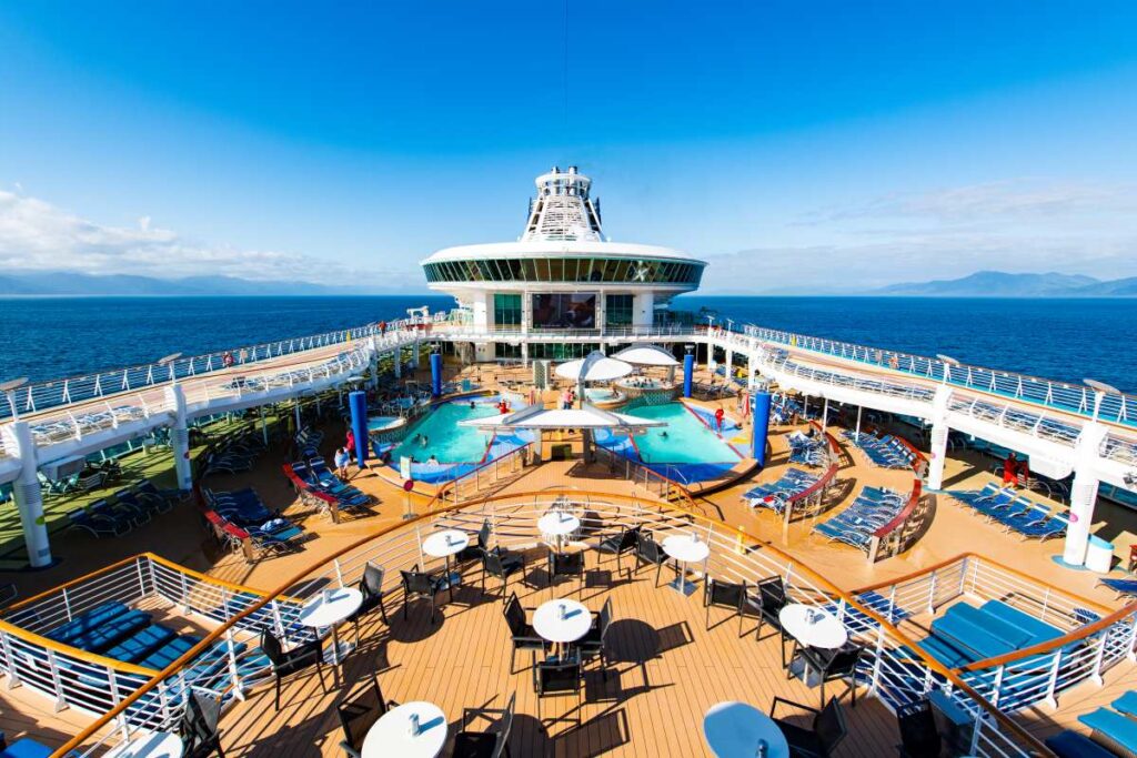 This image showcases the pool deck aboard the Explorer of the Seas, part of the Royal Caribbean fleet. The deck features multiple pools, sun loungers, and shaded seating areas, offering ample space for relaxation under the clear blue sky. The ship's modern design and scenic ocean backdrop create a vibrant and inviting atmosphere for guests to enjoy their cruise experience.
