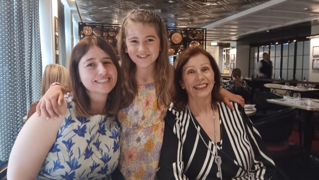 Three generations of a family are smiling and posing together at a dining table in the Le Petit Chef restaurant on the Celebrity Apex cruise ship. The background features elegant decor with a modern design, adding to the cheerful and intimate atmosphere of their dining experience.
