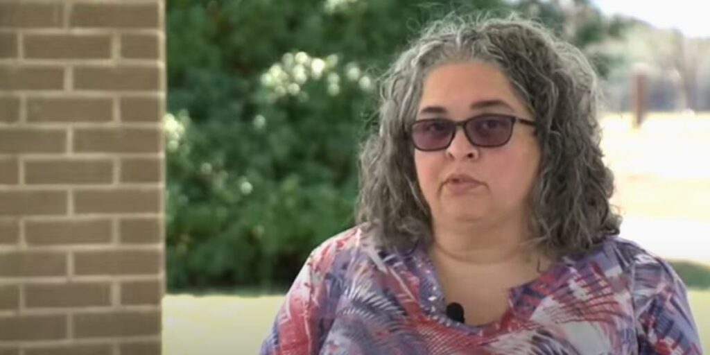 A woman with gray curly hair and sunglasses speaks outdoors near a brick wall and green bushes. She wears a colorful patterned top and has a small microphone clipped near her collar.