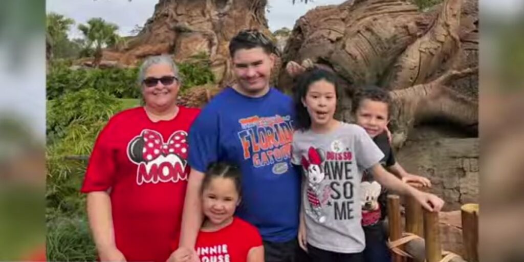 A smiling woman in a red “MOM” Minnie Mouse shirt stands with four children and a man in front of a large tree, possibly at a theme park. The group appears cheerful, with kids wearing Disney-themed shirts.