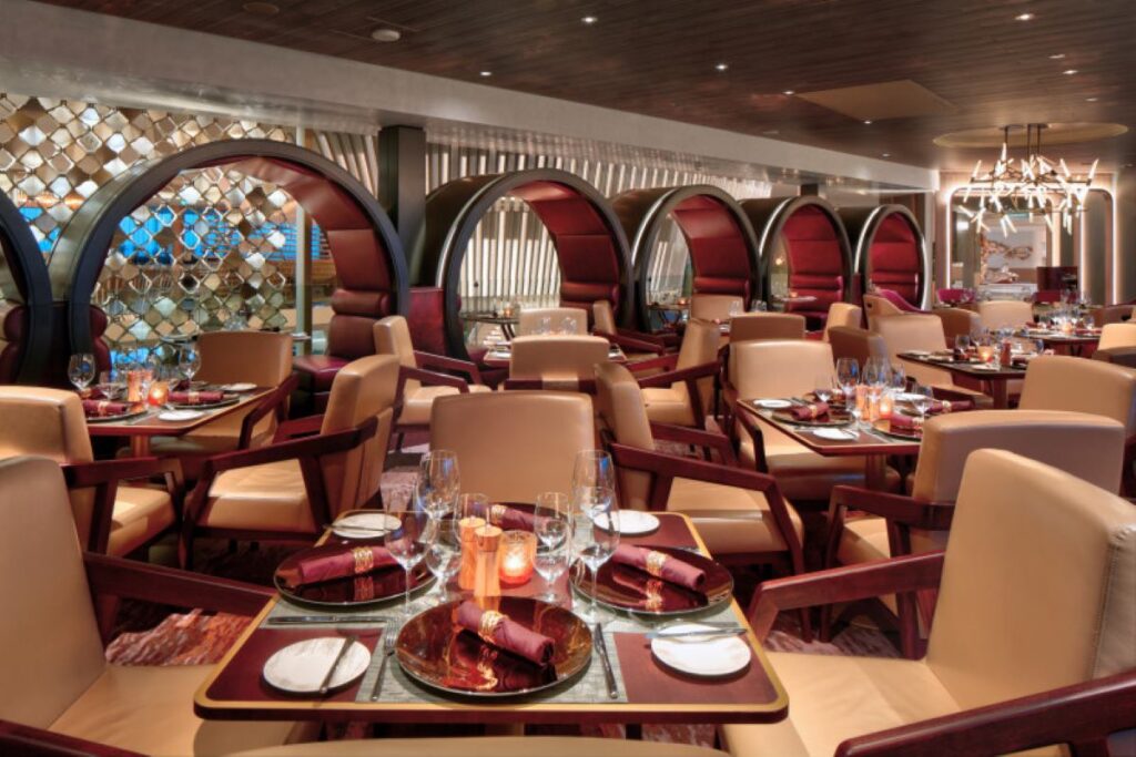 This image showcases the elegant Fine Cut Steakhouse aboard a Celebrity cruise ship, featuring a sophisticated and modern dining space. The restaurant is adorned with luxurious leather seating, rich burgundy accents, and unique arched booths that create an intimate and stylish ambiance. The tables are meticulously set with fine glassware, silverware, and deep red napkins, offering a premium environment for a high-end dining experience.