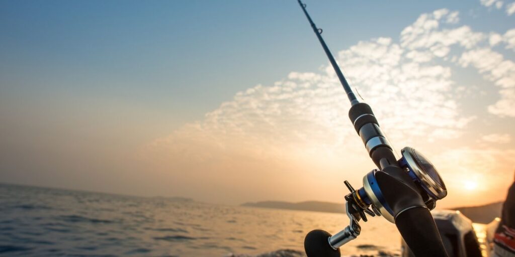 Close-up of a fishing rod angled over the side of a boat at sunset, with calm ocean waters and a soft golden sky. This image represents fishing, an activity typically prohibited directly from cruise ships.