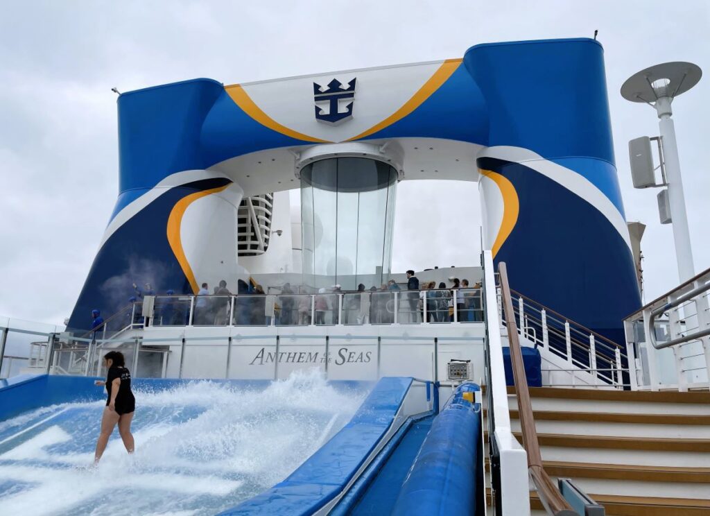 Passenger surfing on the FlowRider surf simulator aboard Royal Caribbean's Anthem of the Seas, with the ship's distinctive blue and yellow funnel in the background.