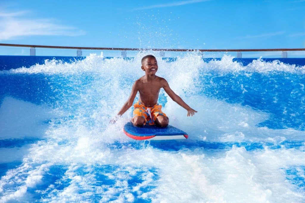 A young boy with a big smile enjoys the exhilarating FlowRider surf simulator on Royal Caribbean's Oasis Class ship, expertly balancing on his board amidst the rushing blue waves.