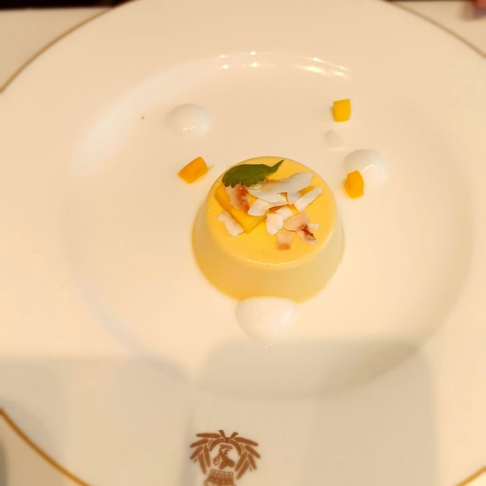 The image shows a beautifully plated dessert from the main dining room on Cunard's Queen Anne cruise ship. The dish features a creamy yellow custard garnished with coconut shavings, small mango cubes, and a sprig of fresh herbs. It is artfully arranged on a white plate with delicate gold trim, displaying the Cunard crest at the bottom, reflecting the ship’s refined dining experience.