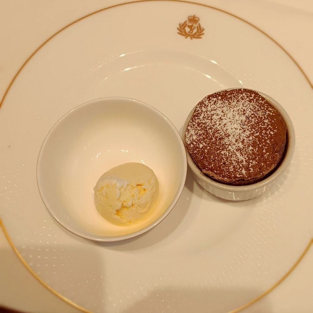 The image shows a dessert from the main dining room on Cunard's Queen Anne cruise ship. A small chocolate soufflé dusted with powdered sugar is served alongside a scoop of vanilla ice cream in a separate bowl. The dish is presented on a fine white plate with a gold trim, featuring Cunard's crest at the top, adding an elegant touch to the gourmet dessert experience.