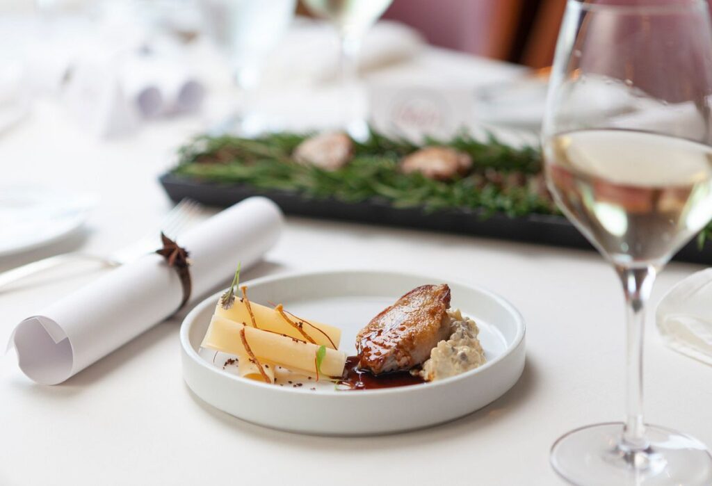 This image captures an exquisite culinary presentation from the Chef's Table on a Carnival Cruise, featuring a beautifully caramelized piece of meat paired with delicate cheese slices and a savory spread. The composition is sophisticated yet inviting, with a glass of white wine in the background that complements the dish, all set against the soft focus of an elegantly laid dining table.