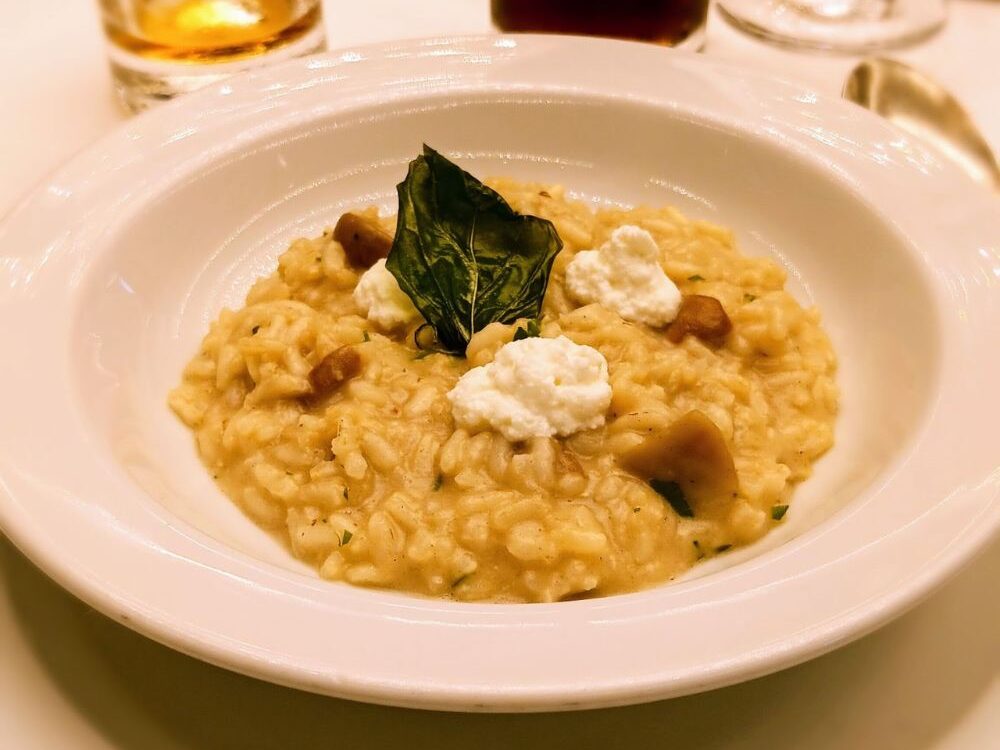 The image shows a creamy risotto dish served in a deep white bowl, topped with small dollops of ricotta cheese and garnished with a crispy basil leaf. The risotto is mixed with pieces of mushrooms, creating a hearty and visually appealing dish, likely enjoyed on an Oceania Cruise ship as part of its fine dining experience. The delicate presentation highlights the gourmet nature of the cuisine served onboard.