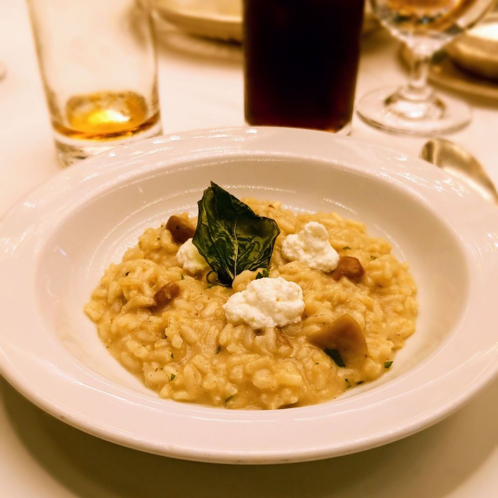 The image shows a creamy risotto dish served in a deep white bowl, topped with small dollops of ricotta cheese and garnished with a crispy basil leaf. The risotto is mixed with pieces of mushrooms, creating a hearty and visually appealing dish, likely enjoyed on an Oceania Cruise ship as part of its fine dining experience. The delicate presentation highlights the gourmet nature of the cuisine served onboard.