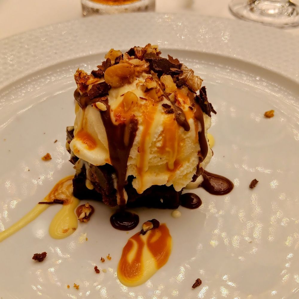 The image showcases a decadent dessert served on an Oceania Cruise ship. It features a scoop of vanilla ice cream atop a rich chocolate brownie, drizzled with caramel and chocolate sauce. The dessert is garnished with chopped nuts and chocolate shavings, offering a delightful mix of textures and flavors. This beautifully presented treat highlights the gourmet culinary experiences provided onboard.