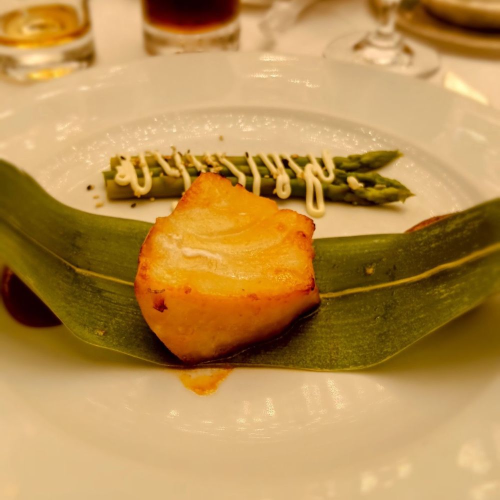 The image shows a fine dining dish consisting of a perfectly seared piece of fish, likely sea bass, presented on a green leaf. In the background, asparagus spears drizzled with a light sauce, likely mayonnaise or aioli, are arranged neatly. The golden, slightly crispy exterior of the fish contrasts beautifully with the fresh green asparagus, highlighting the elegant presentation of food onboard an Oceania Cruise ship.