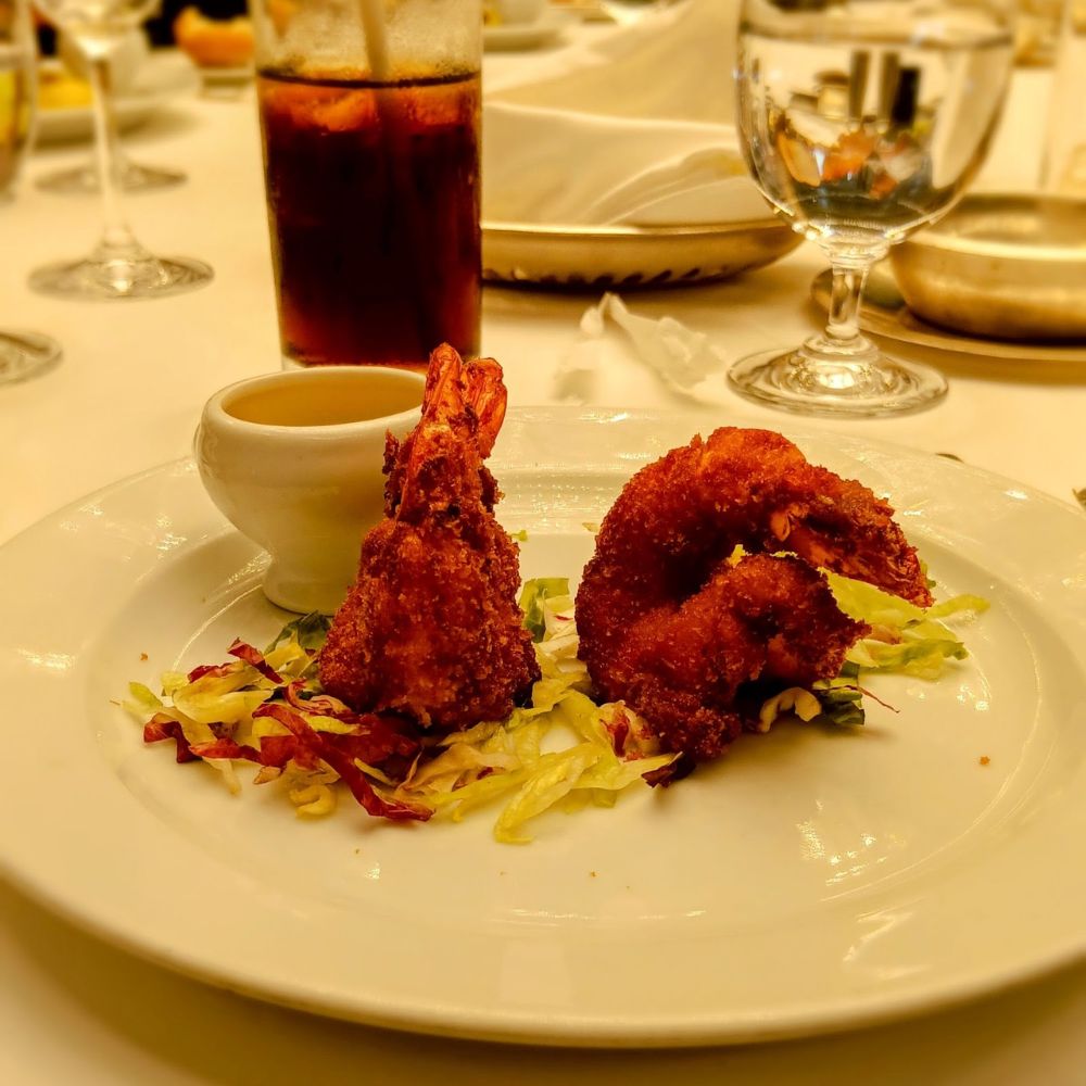 The image shows a beautifully plated dish of breaded jumbo shrimp served on a bed of shredded lettuce and cabbage, accompanied by a small bowl of dipping sauce. The background features a glass of iced beverage and water, with a fine dining setting on an Oceania Cruise ship. The golden-brown crust of the shrimp adds a crispy texture to the dish, aligning with Oceania Cruises' reputation for gourmet dining.