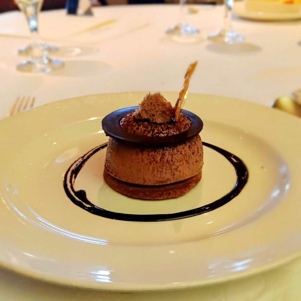 The image displays an elegantly plated dessert served on the Regal Princess cruise ship. The dessert appears to be a chocolate mousse or ganache topped with a decorative chocolate disk and a small garnish. It's set on a thin crust base, with a drizzle of chocolate sauce artfully surrounding it on the plate, making it a visually appealing gourmet treat.