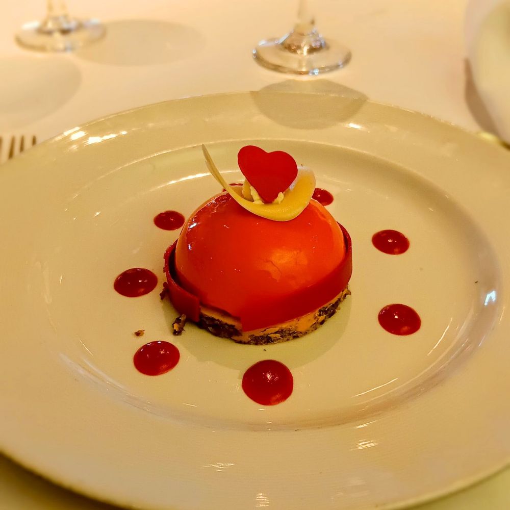 The image features a beautifully plated dessert served on the Regal Princess cruise ship. The dessert is a dome-shaped treat, glazed in a glossy orange-red finish, and decorated with a small red heart and a white chocolate swirl. It sits on a delicate base, surrounded by a circle of neatly placed red sauce dots, likely adding a fruity or tangy flavor contrast to the dish. The presentation is elegant and artistic, perfect for a fine dining experience.