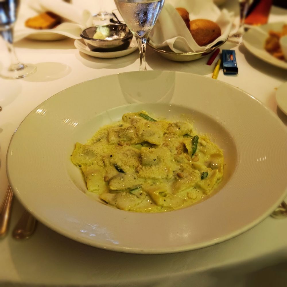 The image shows a dish of pasta, likely ravioli, served on the Regal Princess. The pasta is topped with a creamy sauce and garnished with grated cheese, possibly Parmesan, and pine nuts. The table setting includes a glass of water, bread rolls, and butter in the background, creating a refined dining experience.