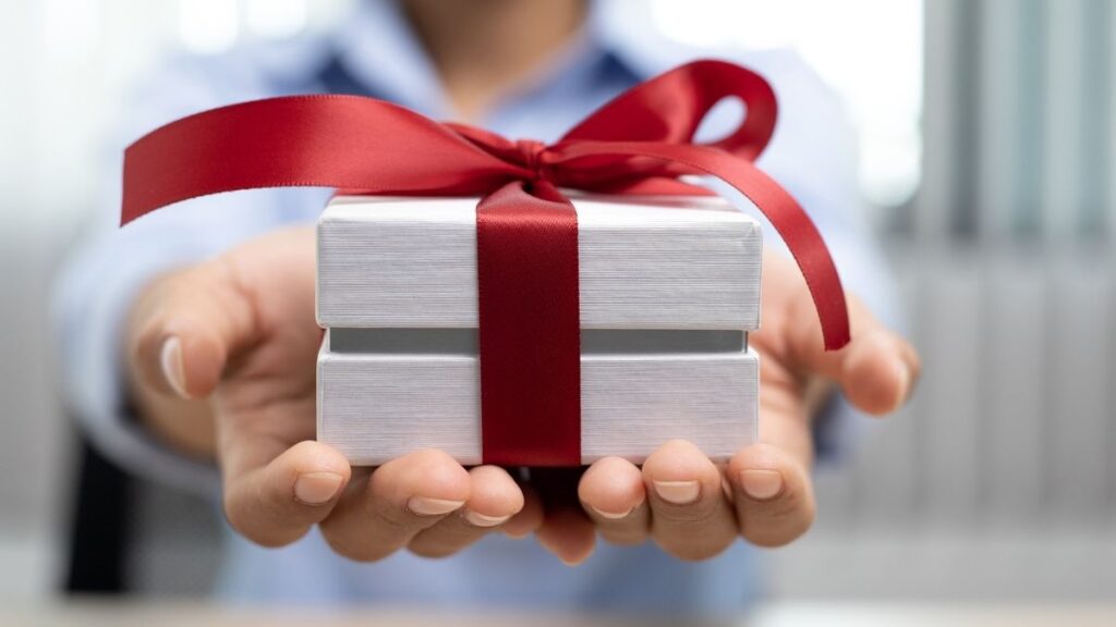 A person wearing a light blue shirt extends their hands, offering a small white gift box wrapped with a neatly tied red ribbon. The background is blurred, emphasizing the present as the main focus. The gesture conveys generosity, celebration, or a special occasion.