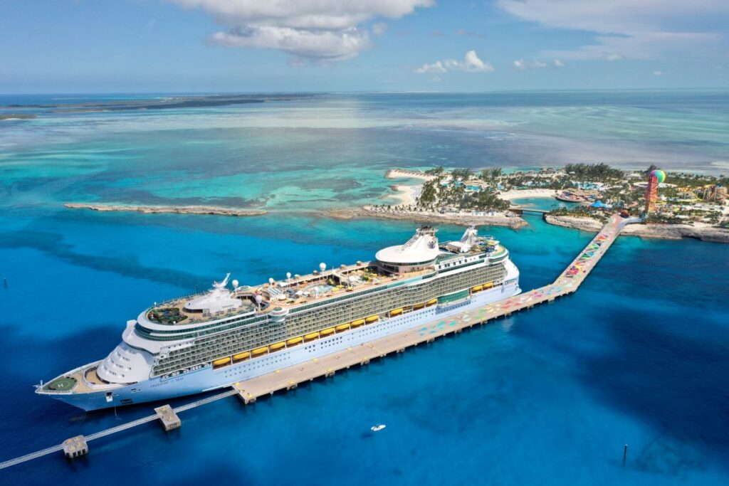 Royal Caribbean's Freedom of the Seas cruise ship is docked at a tropical island, captured from an aerial view. The turquoise and shallow waters of the Caribbean Sea contrast with the large, elegant white ship, while the vibrant island in the background adds to the paradise-like setting.