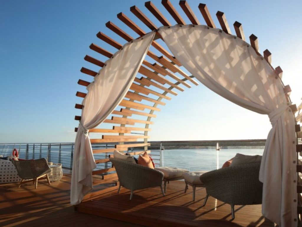 This image showcases the Galapagos Glamping experience on the Celebrity Flora, featuring a luxurious, semi-enclosed wooden cabana with flowing white curtains. Two cozy lounge chairs are arranged for relaxation, overlooking the ocean under a clear sky. The setup provides an inviting, tranquil atmosphere for enjoying the beautiful seascape in comfort and style.