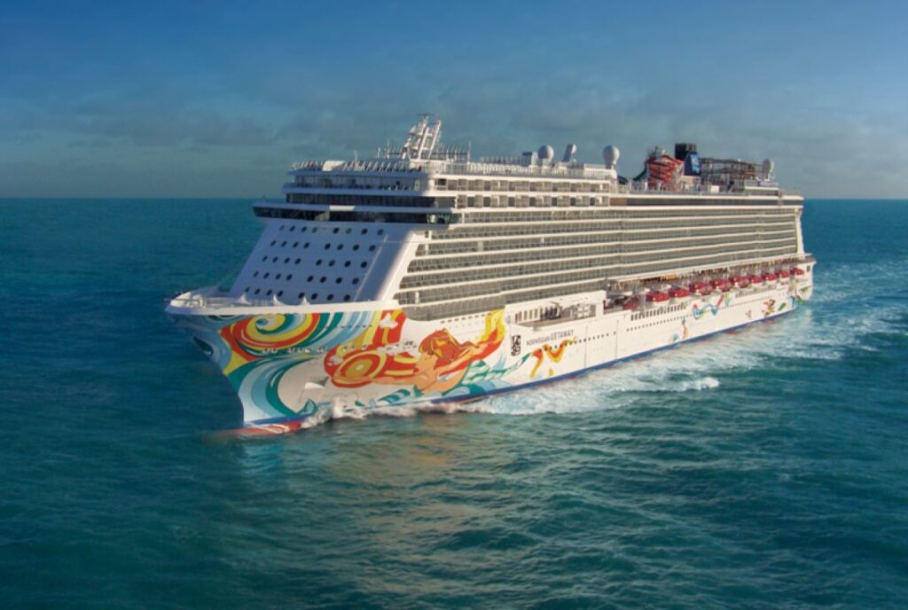 This image shows the Norwegian Getaway cruise ship sailing through turquoise waters under a partially cloudy sky. The ship's hull is adorned with vibrant tropical-themed artwork, reflecting the lively and luxurious experience it offers. Its sleek and expansive design stands out against the calm, open ocean, emphasizing its grandeur.