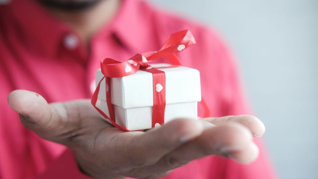 A person holding out their hand with a small gift box, wrapped with red ribbon.