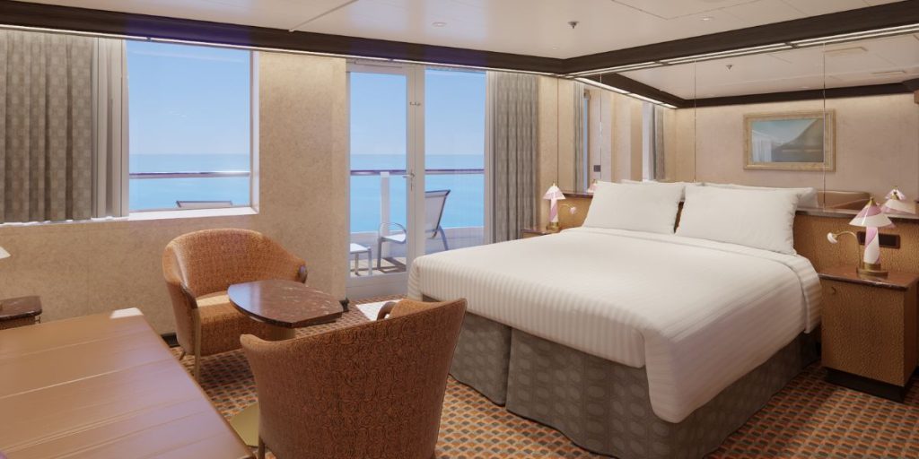 A spacious grand suite with a large bed adorned with crisp white linens, flanked by pink-shaded lamps, and a sitting area with plush chairs. The room boasts a panoramic ocean view through floor-to-ceiling windows that open to a private balcony, inviting natural light and offering a glimpse of the horizon. The elegant decor and generous amenities suggest a luxurious stay aboard the cruise ship.