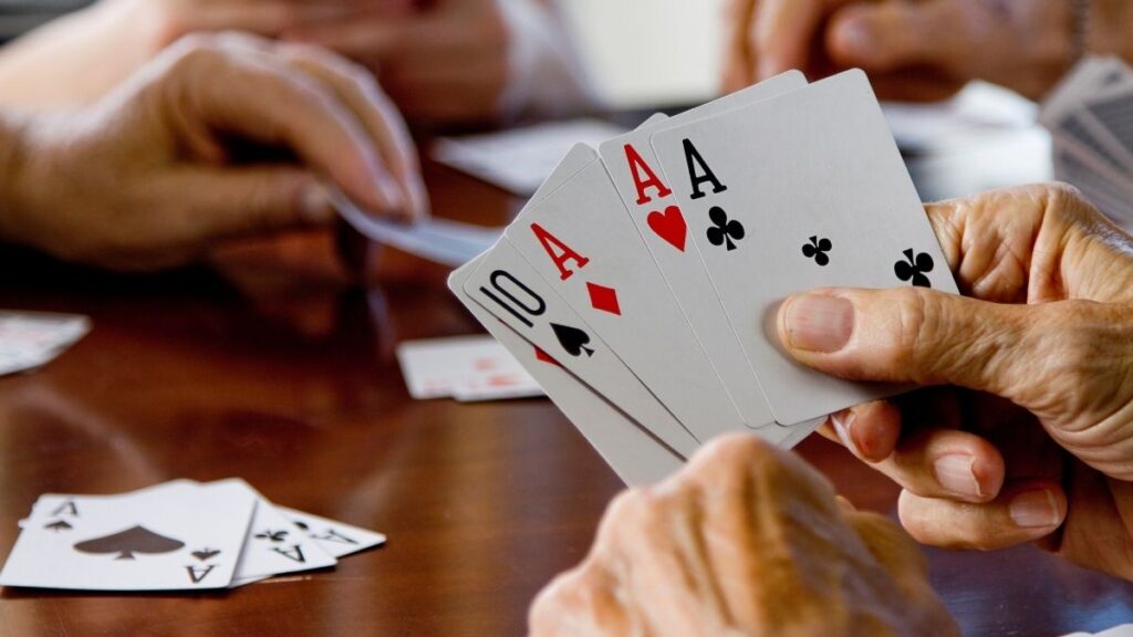 This image shows a close-up of a group of people playing cards, with one person holding a hand of cards featuring three aces and a ten. The wooden table and scattered cards suggest a casual and friendly game in progress.