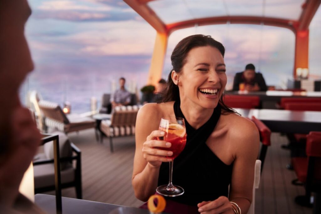 This image features a smiling guest aboard a Celebrity cruise ship, enjoying a vibrant cocktail while seated in an elegant outdoor dining area. The backdrop includes stunning sunset hues and a relaxed atmosphere, with other guests visible in the background. The scene captures the joy and sophistication of the onboard social and dining experience.