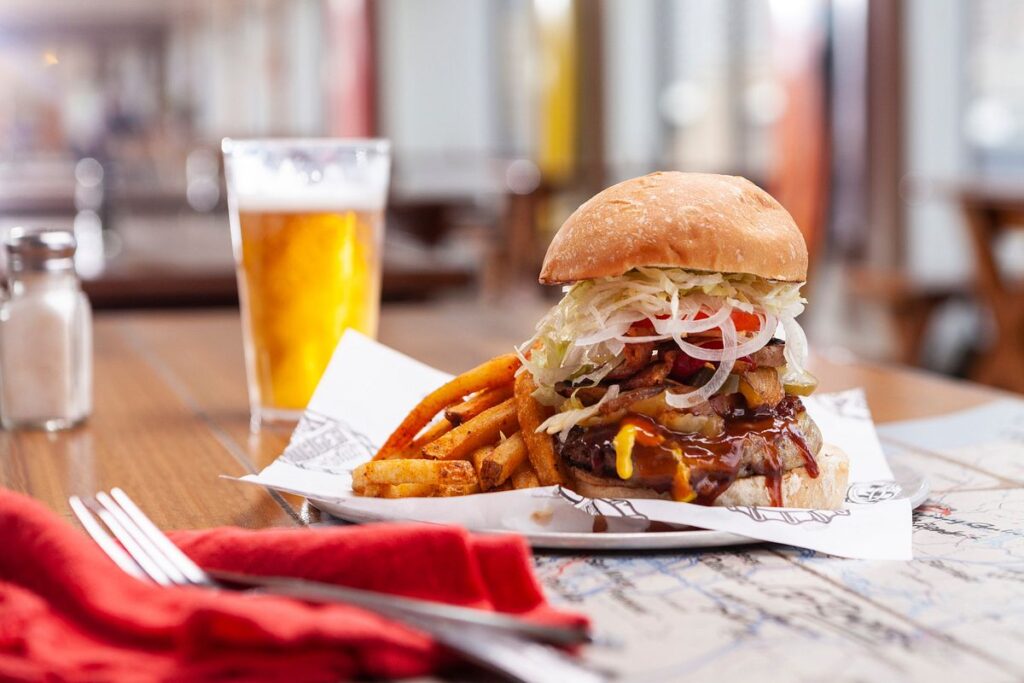 A mouth-watering burger from Guy's Burger Joint on Carnival Spirit, served with seasoned fries and a cold beer, ready to be enjoyed in a casual dining setting.