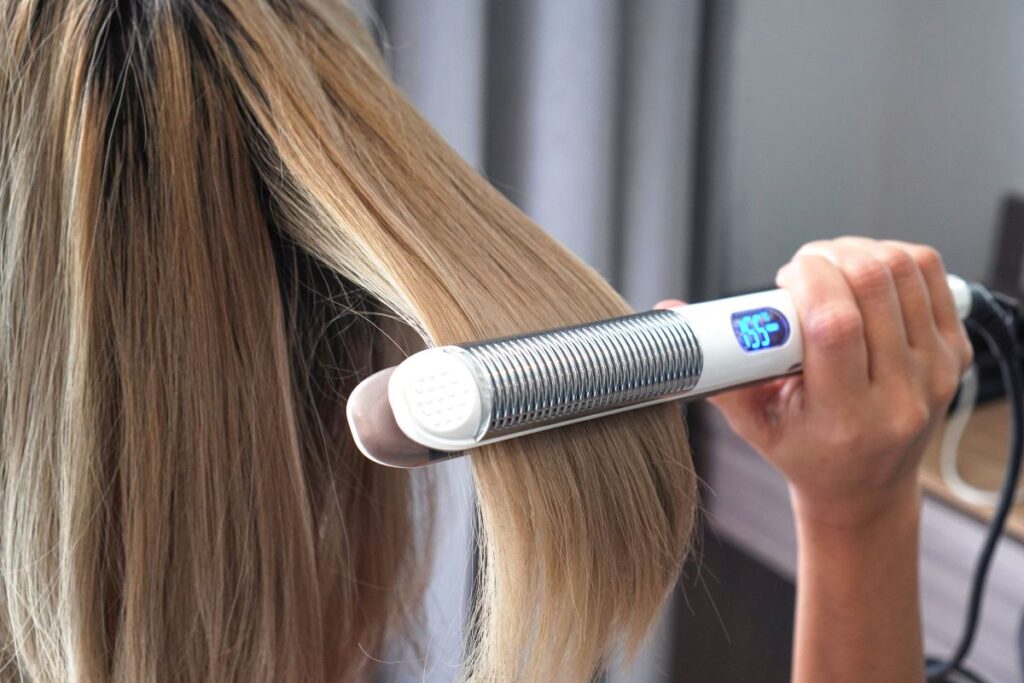 The image depicts a person using a hair straightener on long blonde hair. The straightener displays a digital temperature reading on its screen, suggesting a modern device with adjustable heat settings. The background is softly blurred, emphasizing the hair and styling tool.