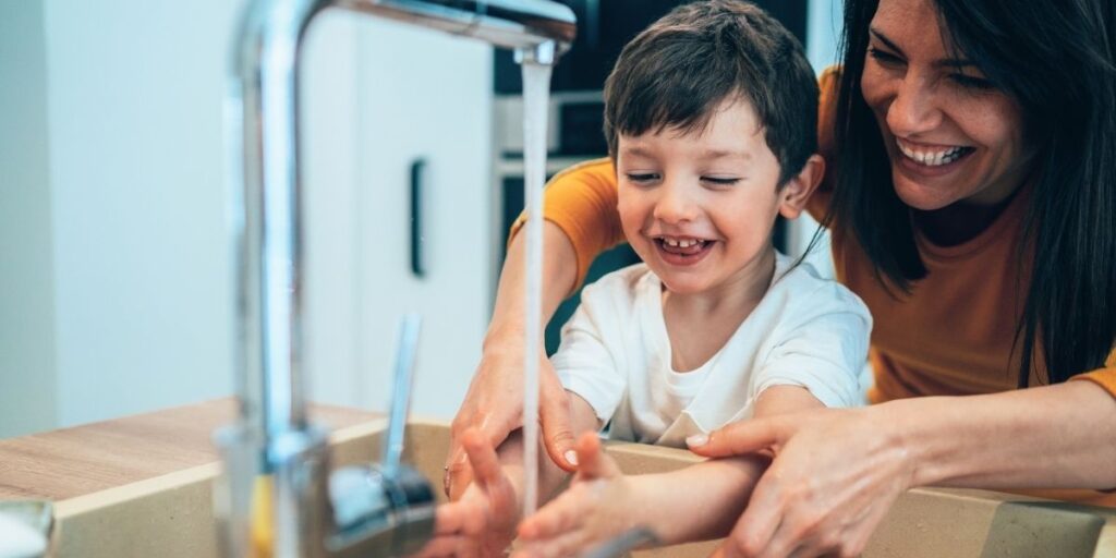 A smiling mother helps her young son wash his hands under a running kitchen faucet, both laughing and enjoying the moment together. The image emphasizes hygiene and bonding in a home setting.