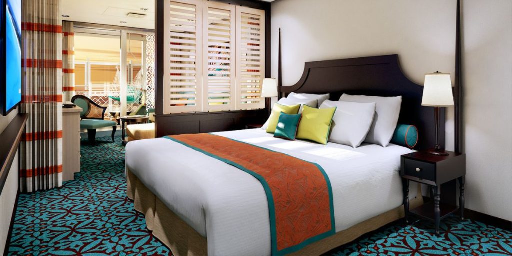 A Havana Cabana Suite, elegantly furnished with a large bed with a crisp white duvet, accented by a vibrant orange and turquoise throw. The room features a tropical-inspired decor, with a stylish chair and access to a private patio area visible through plantation shutters, offering a blend of comfort and exotic flair. The patterned carpet adds a splash of color, complementing the room's warm and inviting ambiance.