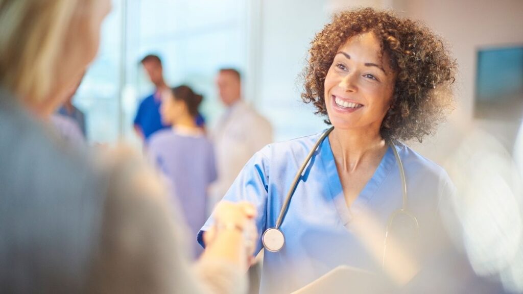 A smiling healthcare professional in blue scrubs with a stethoscope around her neck warmly shaking hands with a patient in a bright, modern medical setting. Other healthcare staff in scrubs and lab coats are blurred in the background, creating a professional yet welcoming atmosphere.