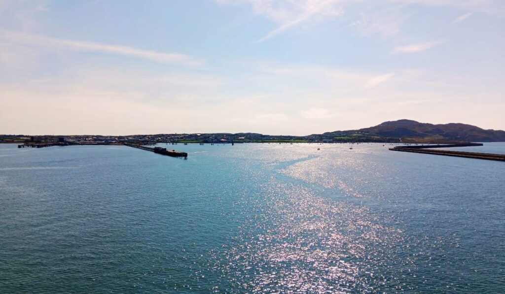The image shows a tranquil view of Holyhead in North Wales on a sunny day. The calm waters of the harbor shimmer in the sunlight, while the coastline, dotted with low buildings, stretches out in the distance. A hilly landscape forms the backdrop, adding to the scenic coastal beauty of the region.