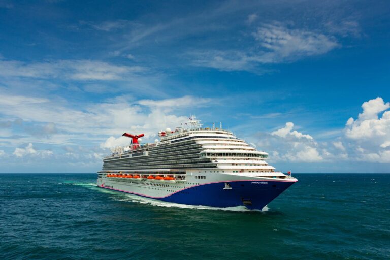 The Carnival Horizon cruise ship glides through turquoise waters, its blue hull complementing the sunny sky dotted with fluffy white clouds above.
