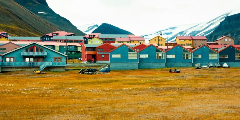 rightly painted houses in shades of blue, red, yellow, and green line a grassy tundra in Svalbard, with snow-capped mountains rising in the background. Snowmobiles and outdoor gear are scattered in front of the colorful homes under an overcast Arctic sky.